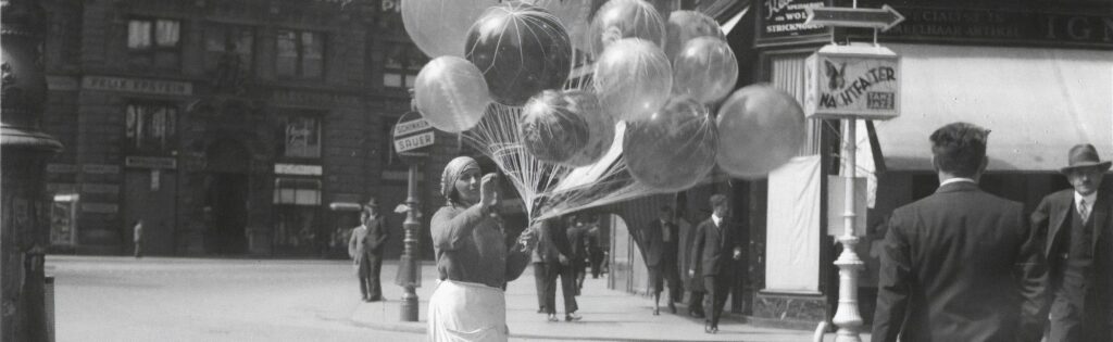 Uma rua do início do século XX. No centro há uma mulher com um lenço na cabeça a vender balões a hélio. 